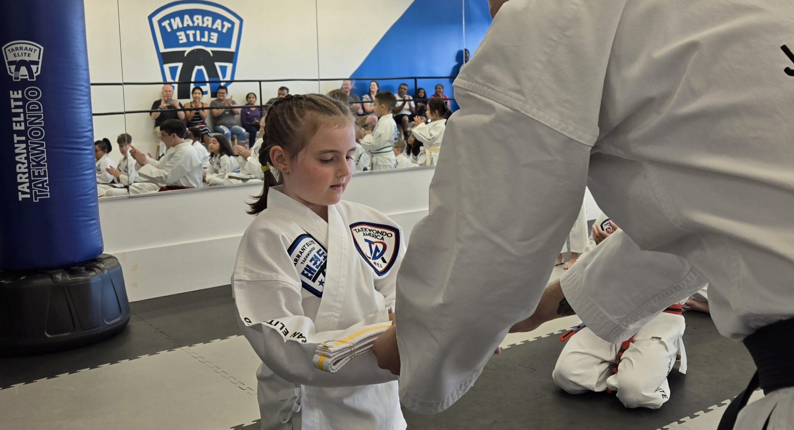 Young girl in a white taekwondo uniform practicing a kick with a partner nearby in a gym.