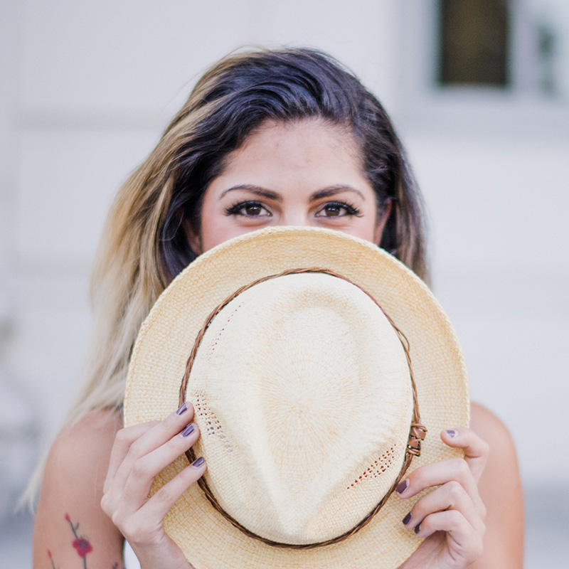 product-fashion-07 › Tarrant Elite Taekwondo Woman with dark and blonde hair holding a straw hat in front of her face, eyes visible over the hat.