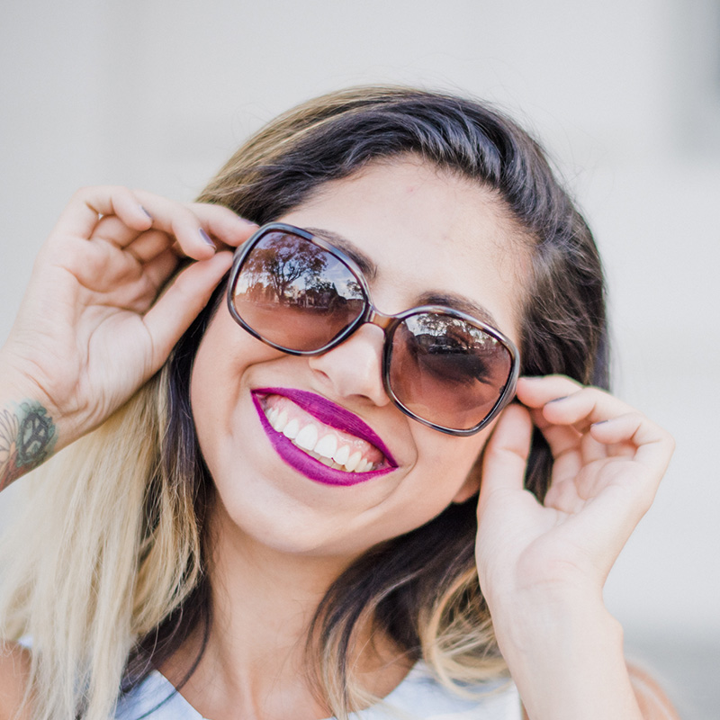 product-fashion-08 › Tarrant Elite Taekwondo Woman with long hair wearing large sunglasses, smiling broadly while adjusting the glasses on her face